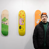 Roméo Elvis stands in front of his exclusive collaboration with THE SKATEROOM, showcasing a collection of limited-edition skate decks featuring his works at the Affordable Art Fair Brussels. The wall behind him displays the artist's name and THE SKATEROOM logo, with vibrant skateboard designs mounted on the wall.