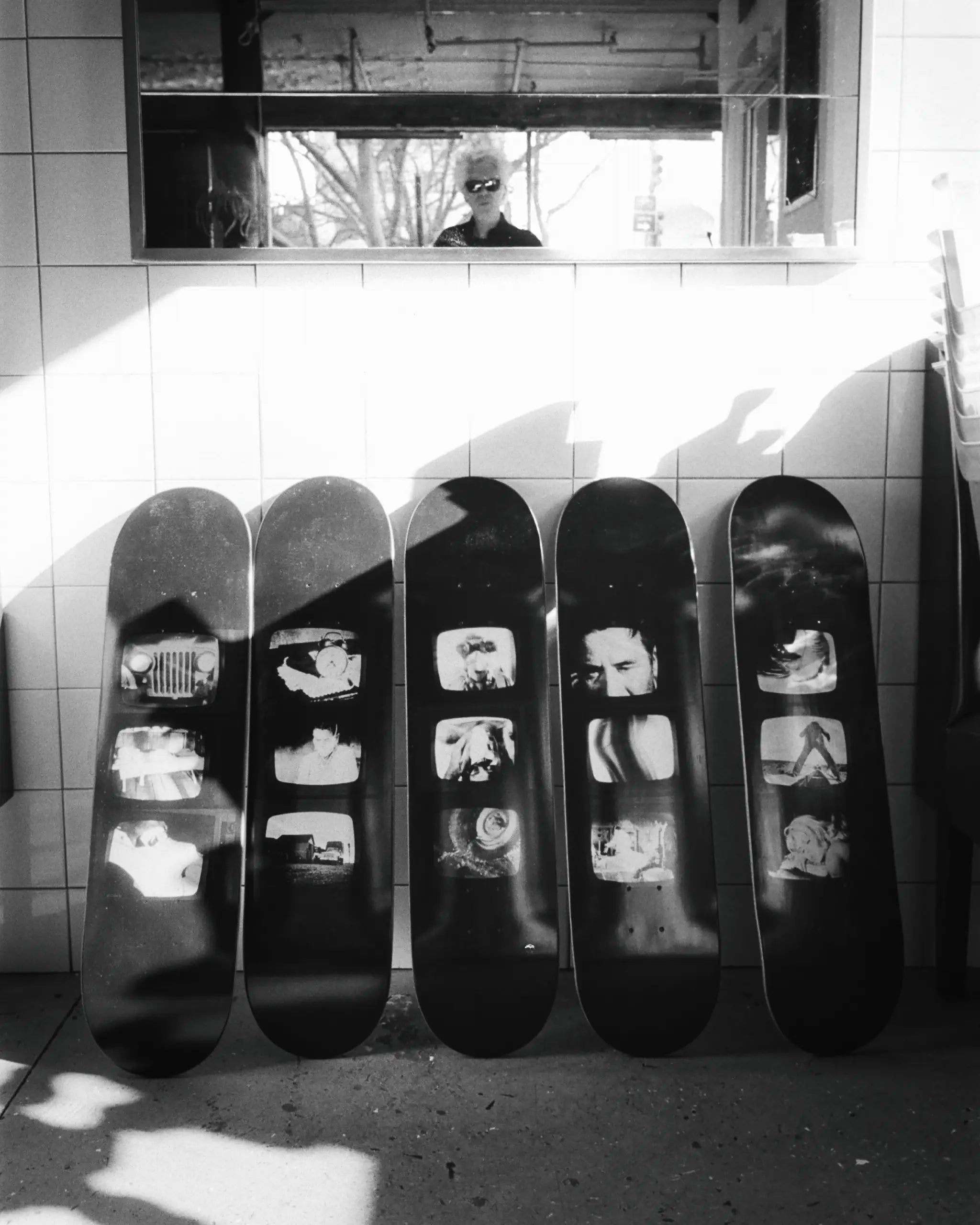 Jim Jarmusch’s ‘TV Photos’ skate deck collection displayed in a striking black-and-white composition. The legendary filmmaker is reflected in the mirror above, adding a cinematic touch to the scene.