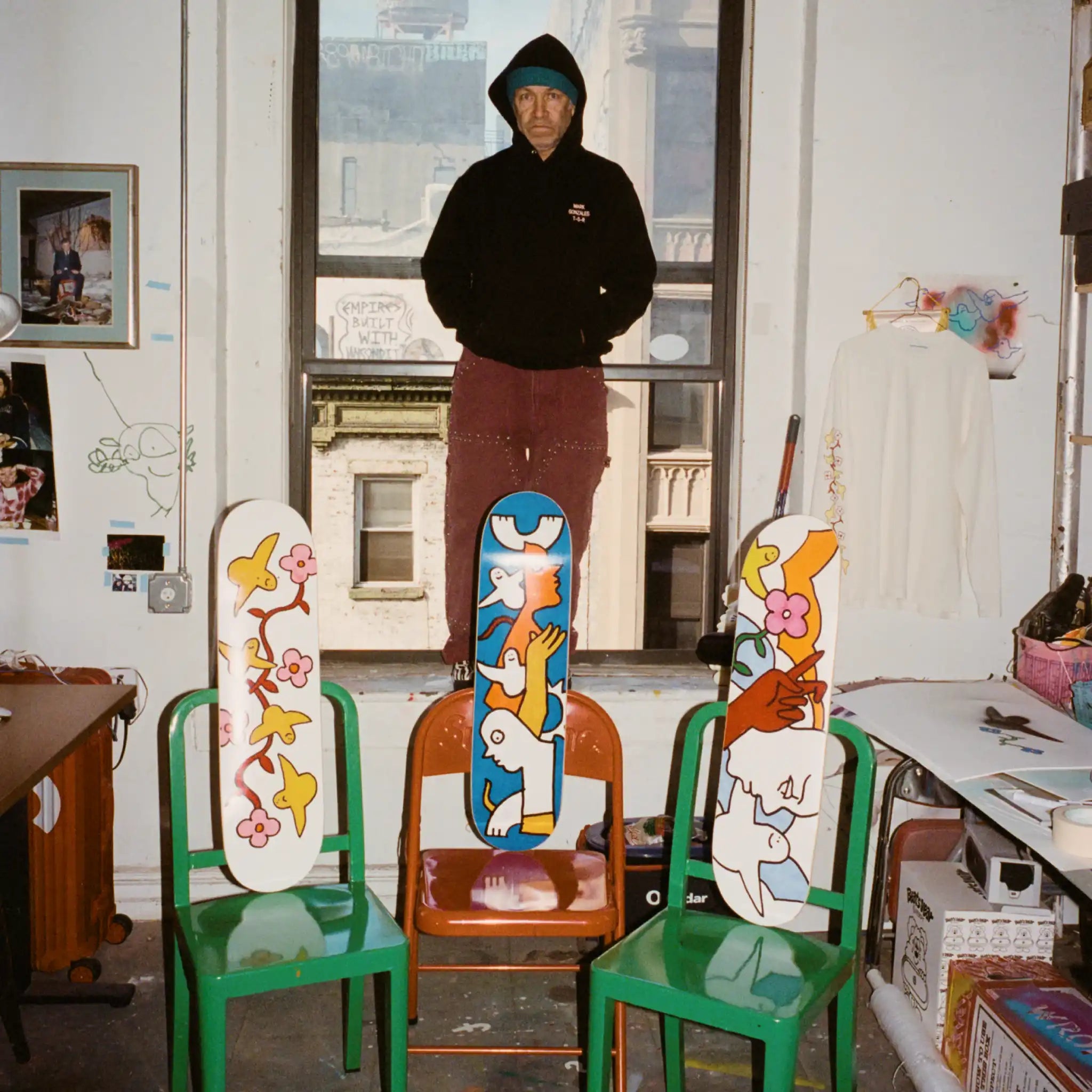 Mark Gonzales standing in his New York studio, surrounded by his signature artwork. Three skate decks featuring his playful, hand-drawn figures and vibrant colors rest on chairs, set against a creative workspace with city views.