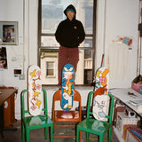Mark Gonzales standing in his New York studio, surrounded by his signature artwork. Three skate decks featuring his playful, hand-drawn figures and vibrant colors rest on chairs, set against a creative workspace with city views.