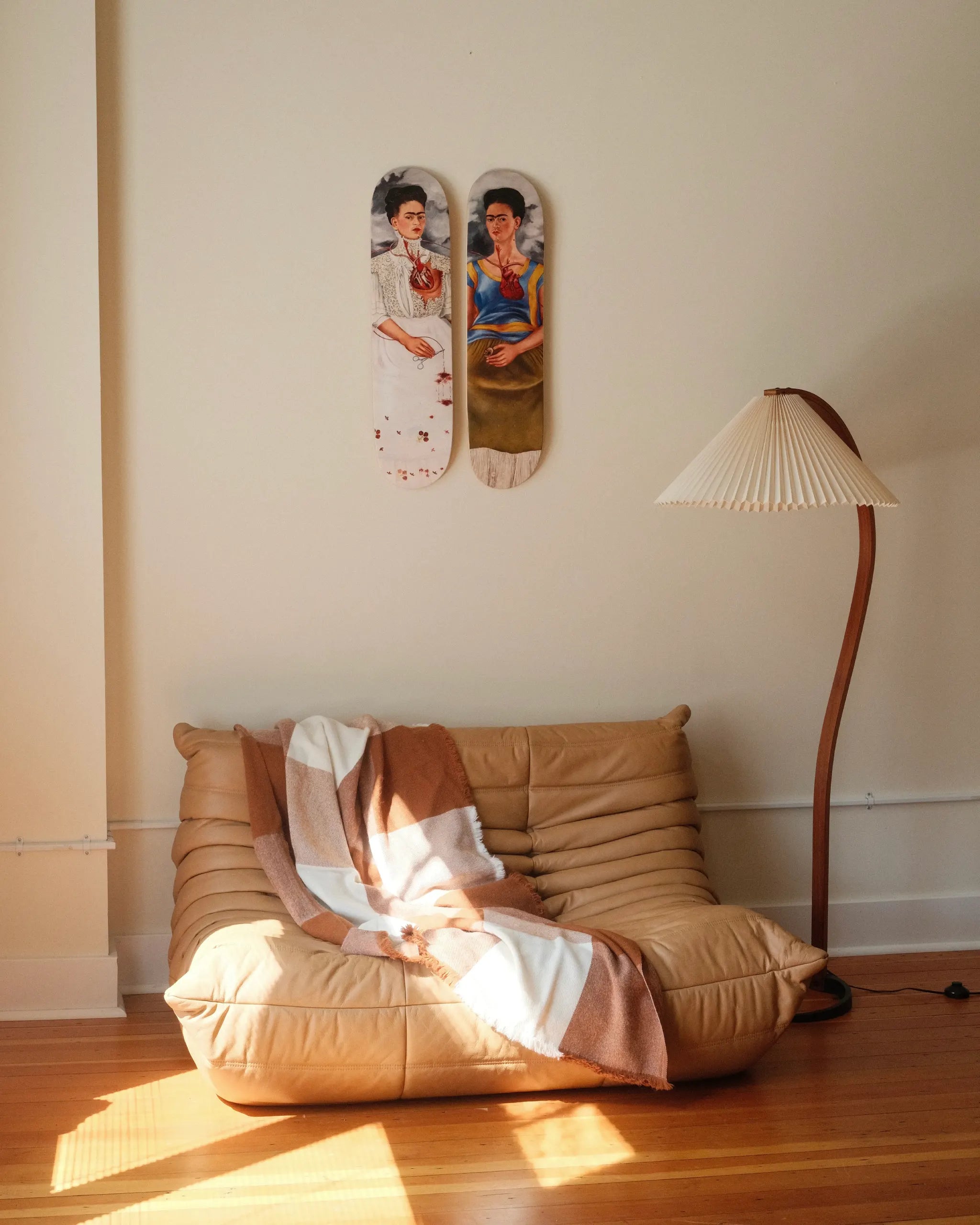 Two Frida Kahlo skateboard decks from THE SKATEROOM’s “The Two Fridas” edition hang above a tan Tog sofa, draped with a checkered throw blanket. A sculptural wooden floor lamp completes the cozy, artful interior.