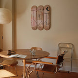 Jean-Michel Basquiat's “Both Poles” triptych skateboard art edition by THE SKATEROOM is displayed above a round wooden table with mid-century modern chairs. A paper lantern and warm afternoon light enhance the minimalist, artful atmosphere.