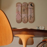 A close-up view of THE SKATEROOM’s “Both Poles” triptych skateboard art edition by Basquiat, mounted on a wall above a tulip-style dining table. The artwork’s hand-drawn elements and soft pink tones contrast with the warm wood and modernist design details in the foreground.
