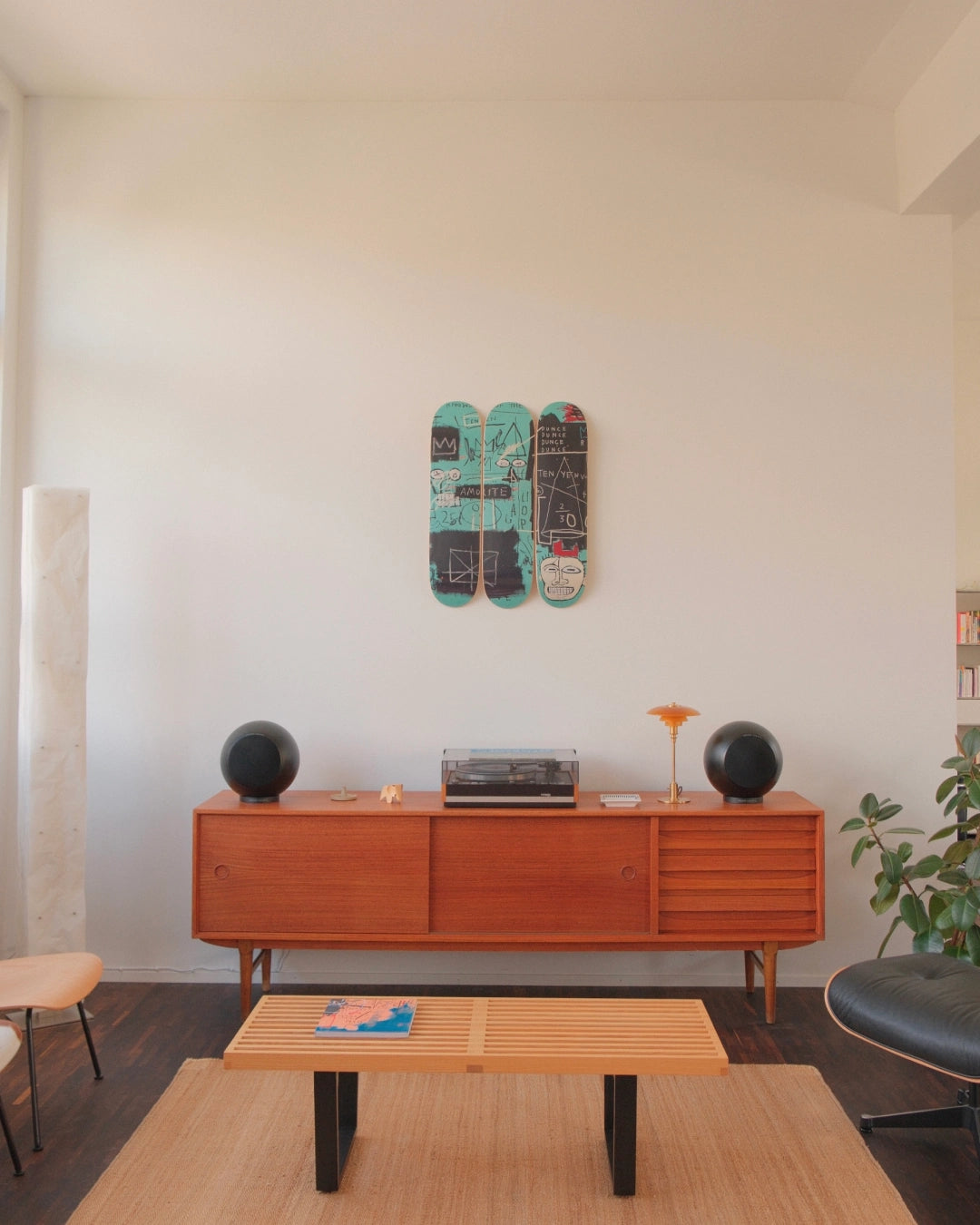 A sun-drenched living space styled with warm modernism and cultural edge. On a crisp white wall, THE SKATEROOM’s Equals Pi triptych by Jean-Michel Basquiat commands attention—its layered graffiti strokes and coded chaos vibrating against the calm. Below, a teak credenza hosts a vintage turntable and amber-lit lamp, flanked by sculptural black speakers. The room is anchored by an Eames lounge chair, boucle beanbag, and design objects that balance soft tactility with mid-century structure.