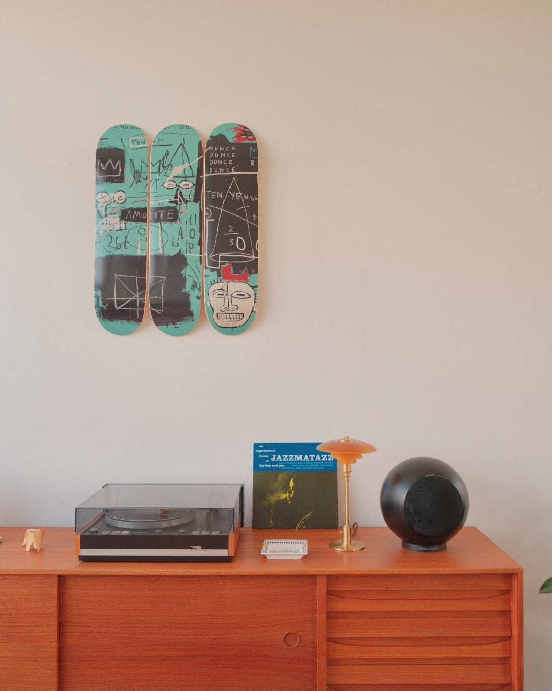Close-up view of Jean-Michel Basquiat’s Equals Pi triptych from THE SKATEROOM, wall-mounted above a warm-toned mid-century sideboard. The skateboards crackle with Basquiat’s signature glyphs—crowns, faces, equations—rendered in teal, black, and chalk-white strokes. Below, a sleek turntable, a Guru “Jazzmatazz” LP, and sculptural speakers anchor the scene in analog cool. An amber lamp glows softly, adding a cinematic warmth.