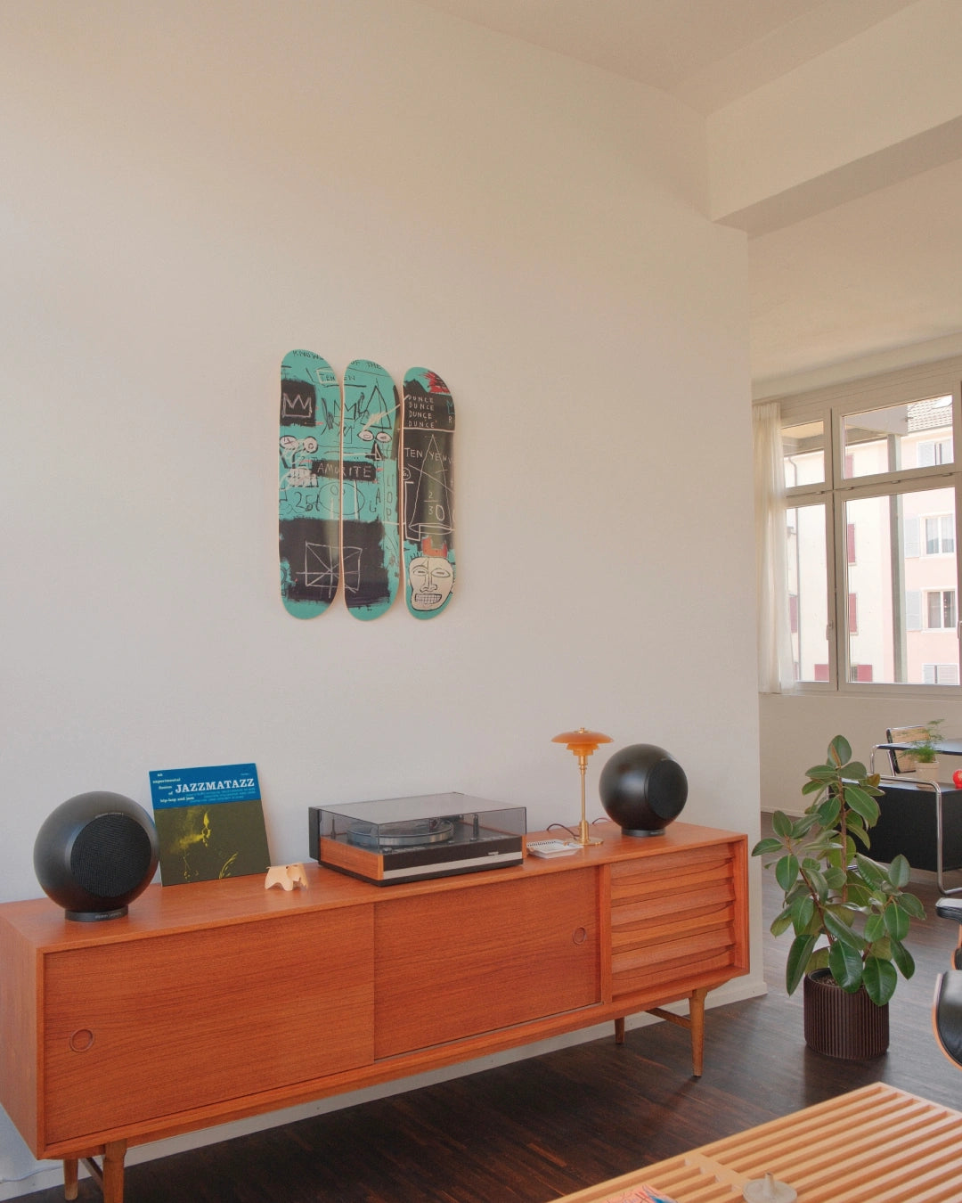 Photograph of the Equals Pi Basquiat triptych skateboard art edition, displayed vertically in a softly lit, minimalist interior. The decks lean against a pale concrete wall, casting subtle shadows. The iconic Basquiat crown and scrawled anatomical symbols pulse with raw energy across the decks. Nearby, a vintage Eames-style chair and abstract decor elements nod to the tension between street culture and high art.
