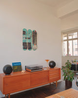 Photograph of the Equals Pi Basquiat triptych skateboard art edition, displayed vertically in a softly lit, minimalist interior. The decks lean against a pale concrete wall, casting subtle shadows. The iconic Basquiat crown and scrawled anatomical symbols pulse with raw energy across the decks. Nearby, a vintage Eames-style chair and abstract decor elements nod to the tension between street culture and high art.