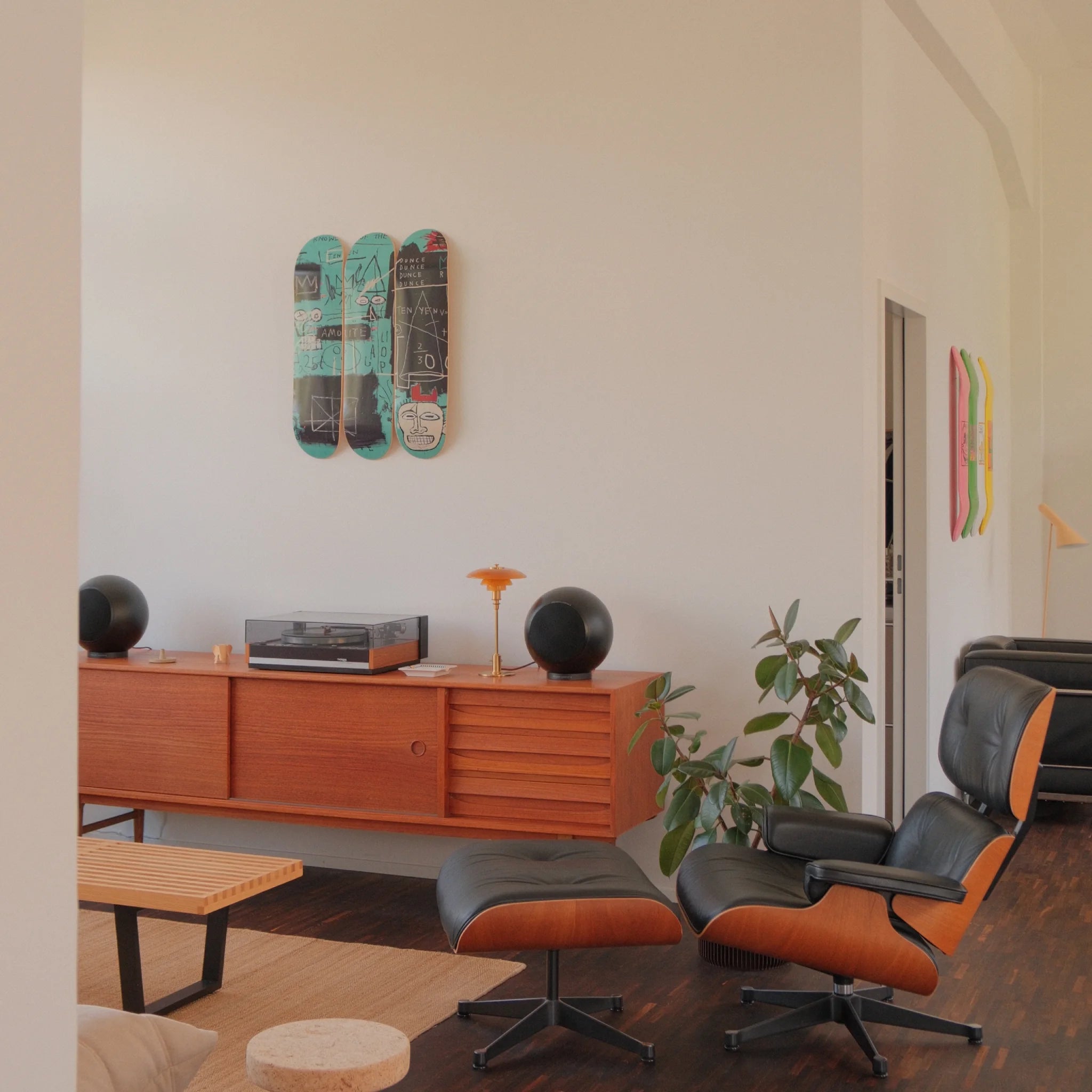 A sun-drenched living space styled with warm modernism and cultural edge. On a crisp white wall, THE SKATEROOM’s Equals Pi triptych by Jean-Michel Basquiat commands attention—its layered graffiti strokes and coded chaos vibrating against the calm. Below, a teak credenza hosts a vintage turntable and amber-lit lamp, flanked by sculptural black speakers. The room is anchored by an Eames lounge chair, boucle beanbag, and design objects that balance soft tactility with mid-century structure.
