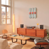 A mid-century modern living room featuring the Jeff Koons "Three Ball 50/50 Tank (Two Spalding Dr. J Silver Series, Wilson Supershot)" triptych skateboard edition mounted on the wall above a wooden sideboard. The space is styled with a turntable, speakers, and a warm ambient lamp, alongside a plush cream-colored sofa and a black Eames lounge chair. A bookshelf and indoor plants complete the refined aesthetic, blending contemporary art with classic design.