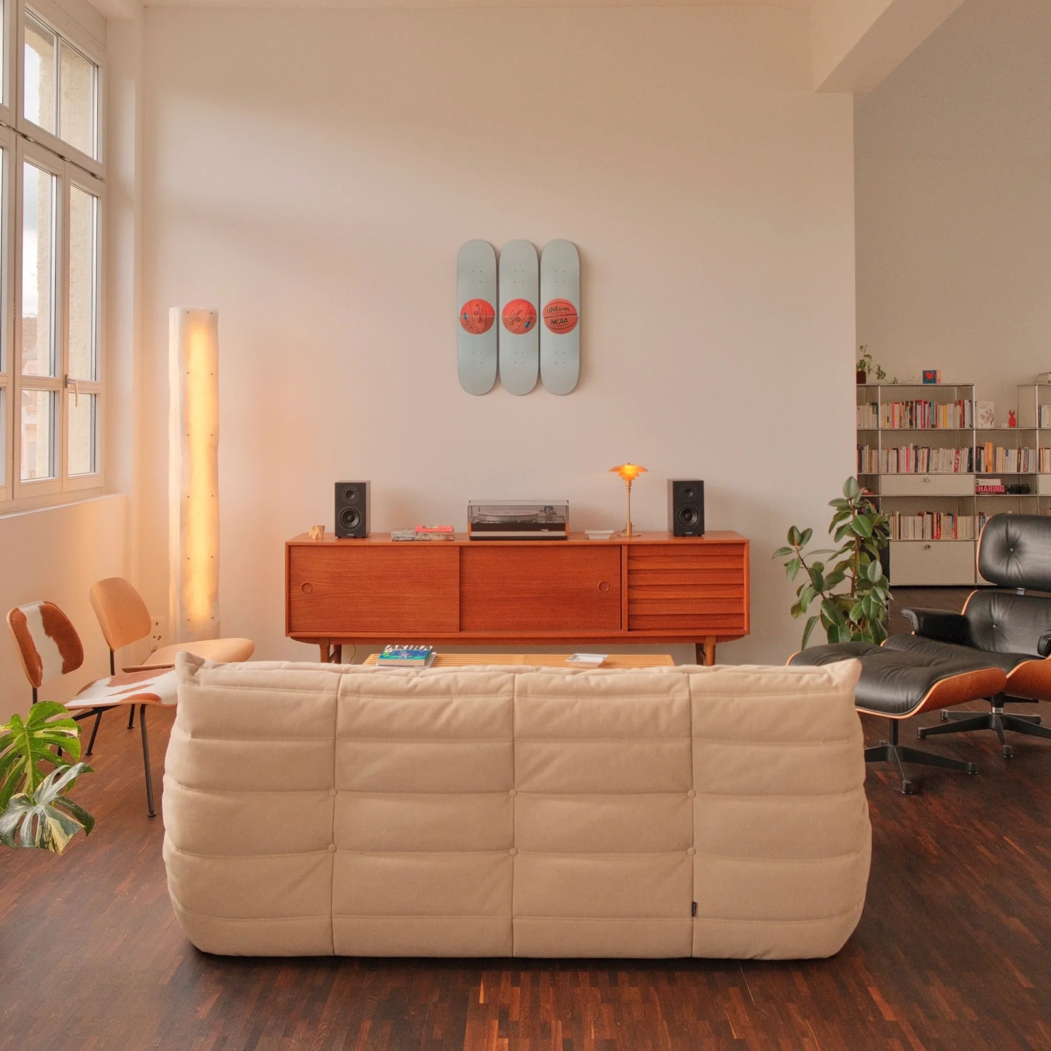 A mid-century modern living room featuring the Jeff Koons "Three Ball 50/50 Tank (Two Spalding Dr. J Silver Series, Wilson Supershot)" triptych skateboard edition mounted on the wall above a wooden sideboard. The space is styled with a turntable, speakers, and a warm ambient lamp, alongside a plush cream-colored sofa and a black Eames lounge chair. A bookshelf and indoor plants complete the refined aesthetic, blending contemporary art with classic design.