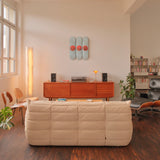 A mid-century modern living room featuring the Jeff Koons "Three Ball 50/50 Tank (Two Spalding Dr. J Silver Series, Wilson Supershot)" triptych skateboard edition mounted on the wall above a wooden sideboard. The space is styled with a turntable, speakers, and a warm ambient lamp, alongside a plush cream-colored sofa and a black Eames lounge chair. A bookshelf and indoor plants complete the refined aesthetic, blending contemporary art with classic design.