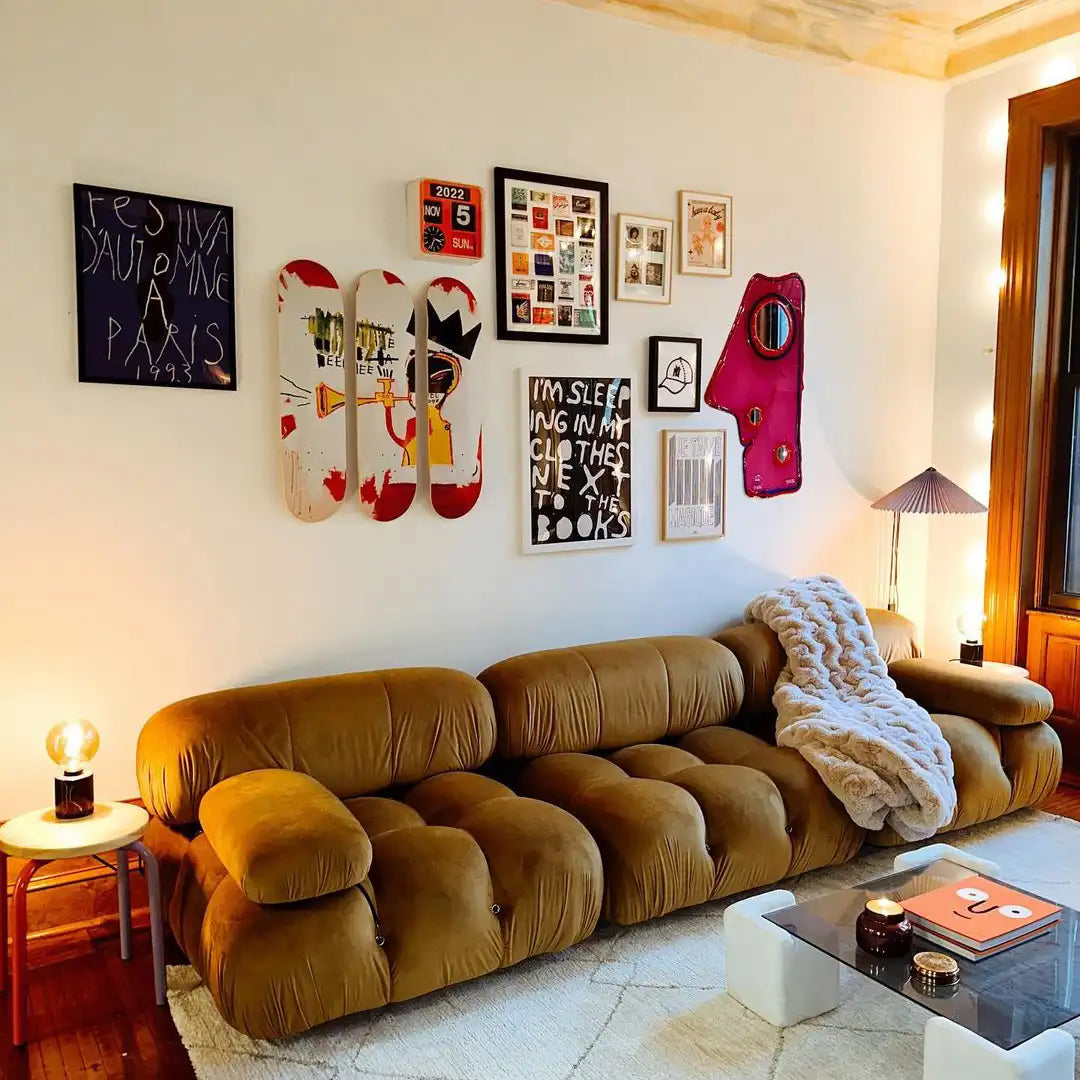 Stylish living room featuring a mustard-toned modular sofa, accented by a gallery wall with eclectic framed art pieces and a Jean-Michel Basquiat "Trumpet" skateboard triptych by THE SKATEROOM, creating a modern and artistic vibe.