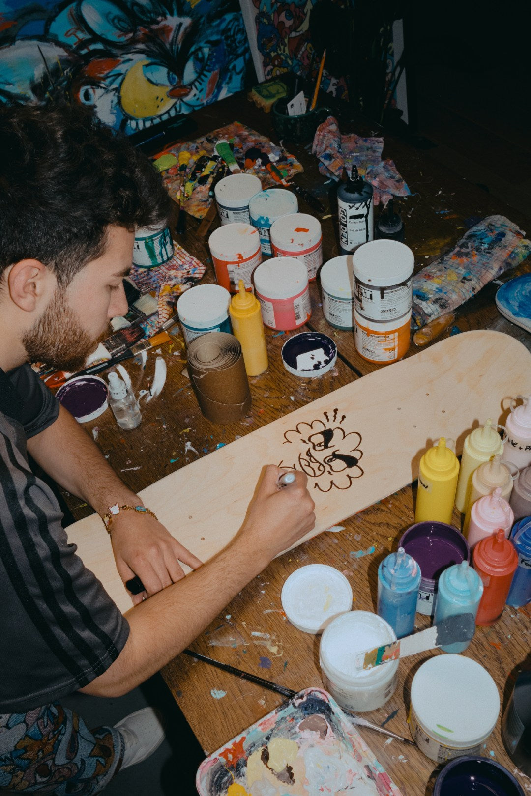 Artist VEXX hand-signing a wooden skate deck with a black marker, drawing a unique doodle character in his paint-covered studio workspace.