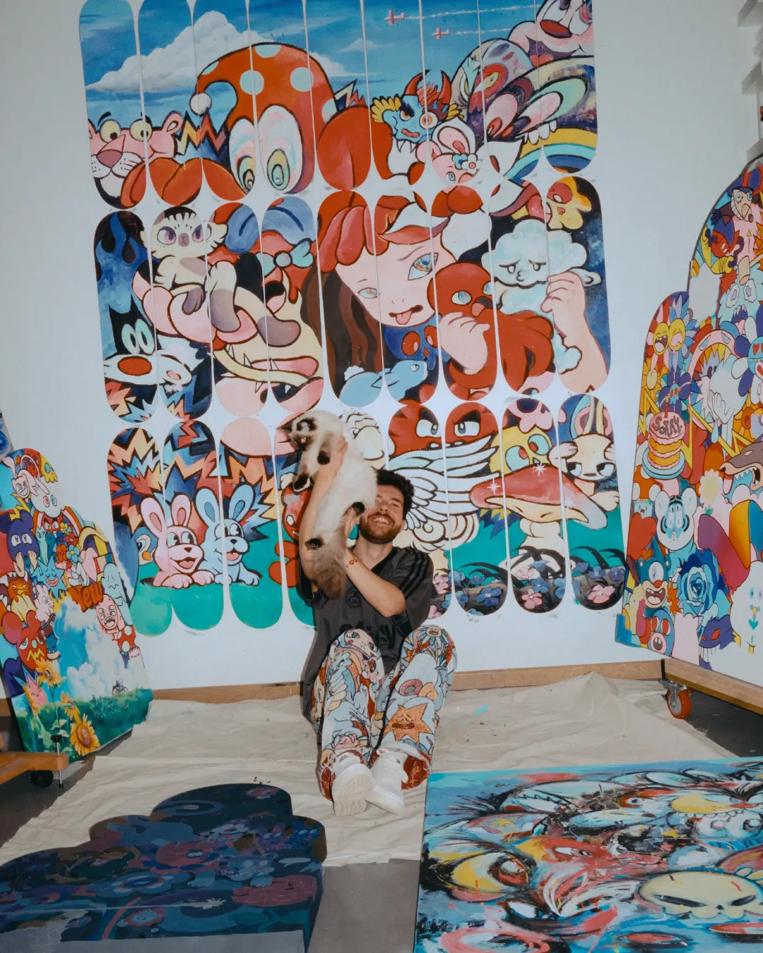 Artist VEXX smiling while holding a cat in front of his completed mural ‘The Life of the Party,’ surrounded by colorful original artworks and painted skate decks in his studio.