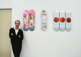 Jeff Koons standing next to his skateboard art editions by THE SKATEROOM featuring 'Pink Panther', 'Three Ball 50/50 Tank (Two Spalding Dr. J Silver Series, Wilson Supershot)' and 'New Shelton Wet/Dry 10 Gallon Doubledecker' designs.