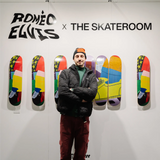 Roméo Elvis stands in front of his exclusive collaboration with THE SKATEROOM, showcasing a collection of limited-edition skate decks featuring his works at the Affordable Art Fair Brussels. The wall behind him displays the artist's name and THE SKATEROOM logo, with vibrant skateboard designs mounted on the wall.