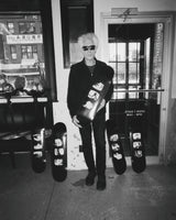 Filmmaker Jim Jarmusch holding one of his limited-edition 'TV Photos' skate decks, standing alongside the full collection. Shot in black and white, the image captures Jarmusch’s signature cool aesthetic, blending film, photography, and skate culture.