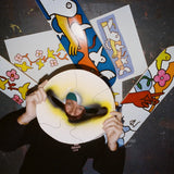 Mark Gonzales lying on the floor, holding a circular artwork with a spray-painted face cutout, partially obscuring his face. Surrounding him are several skateboard decks and art prints from his collaboration with THE SKATEROOM, featuring his signature playful, abstract characters and vibrant designs. The image captures Gonzales' signature artistic style and the essence of the collection.