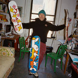 Mark Gonzales standing in his New York studio, holding three skate decks from his collaboration with THE SKATEROOM. Each deck features his signature playful, abstract figures and vibrant colors. The studio space is filled with art supplies, green chairs, and creative energy, reflecting Gonzales' artistic world.