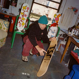 Mark Gonzales in his New York studio, crouching while spray-painting the underside of a blank skate deck with his signature lettering. Behind him, his collaborative skate decks with THE SKATEROOM are displayed on green chairs, featuring his signature playful figures and abstract designs. The workspace is filled with art supplies, paint cans, and creative energy.