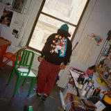 Mark Gonzales standing in his studio wearing a black hoodie from his collaboration with THE SKATEROOM, featuring colorful illustrations on the back. The studio is filled with art supplies, spray cans, and a creative atmosphere, capturing the essence of his artistic space in New York City.