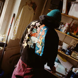 Mark Gonzales in his New York studio, seen from behind wearing a black hoodie featuring his signature artwork in collaboration with THE SKATEROOM. The hoodie showcases his iconic playful figures intertwined with floral elements in vibrant colors. In the background, a long-sleeve shirt from the collection hangs on the wall, while shelves filled with art supplies and sketches reflect his creative workspace.