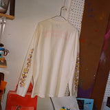 Mark Gonzales x THE SKATEROOM long-sleeve t-shirt hanging in a creative studio. The cream-colored shirt features signature Gonzales artwork with yellow doves and pink flowers running down the sleeves, and subtle embroidered text on the back.