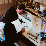 Mark Gonzales seated at a cluttered workspace, sketching a signature artwork featuring a black bird and blue flowers. He wears a black hoodie with his signature colorful character design on the back. The desk is filled with art supplies, sketches, and prints from his collaboration with THE SKATEROOM, capturing the creative process behind the collection.