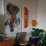 Close-up of a vibrant and modern living space featuring a Warhol Campbell’s Soup Peach skateboard deck mounted on the wall, accompanied by an abstract art print, lush green plants, and a stylish turntable setup with a golden record.