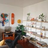 Stylish living room setup featuring a vibrant Warhol Campbell’s Soup Peach skateboard deck mounted on the wall, accompanied by modern art prints, lush green plants, and minimalist shelving adorned with books, decor items, and a turntable for a cozy and creative atmosphere.