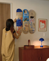 Person arranging Basquiat skate decks on a wall with a lamp and mid-century cabinet in the foreground.