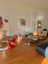 Modern living space featuring a stylish gallery wall with a Jean-Michel Basquiat skateboard triptych by THE SKATEROOM, mid-century furniture, a vibrant red Panton chair, and natural light accentuating the room's minimalist aesthetic.