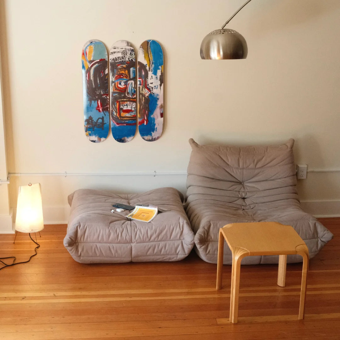 Three Jean-Michel Basquiat "Skull" skate decks on a wall above two Togo sofas with a small wooden table in front.