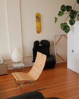 In a softly lit corner, THE SKATEROOM’s Untitled (New York) edition by Jean-Michel Basquiat hangs like a relic—graffiti-scrawled and defiant, in bold yellow against a muted wall. Beneath it, a sculptural black armchair and rattan chaise form a clash of textures: brutalism meets ease. A paper lantern, unframed canvas, and lush monstera leaves round out the scene, evoking the creative quiet of a lived-in studio. It’s a portrait of presence—art not as object, but as atmosphere.