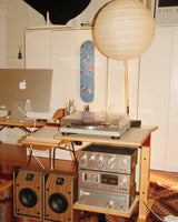 Record player with vintage speakers and a Sky Blue Kandinsky skateboard on a shelf in a room.