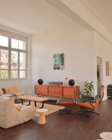 A sun-drenched living space styled with warm modernism and cultural edge. On a crisp white wall, THE SKATEROOM’s Equals Pi triptych by Jean-Michel Basquiat commands attention—its layered graffiti strokes and coded chaos vibrating against the calm. Below, a teak credenza hosts a vintage turntable and amber-lit lamp, flanked by sculptural black speakers. The room is anchored by an Eames lounge chair, boucle beanbag, and design objects that balance soft tactility with mid-century structure. 