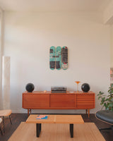 A sun-drenched living space styled with warm modernism and cultural edge. On a crisp white wall, THE SKATEROOM’s Equals Pi triptych by Jean-Michel Basquiat commands attention—its layered graffiti strokes and coded chaos vibrating against the calm. Below, a teak credenza hosts a vintage turntable and amber-lit lamp, flanked by sculptural black speakers. The room is anchored by an Eames lounge chair, boucle beanbag, and design objects that balance soft tactility with mid-century structure. 