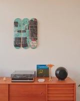 Close-up view of Jean-Michel Basquiat’s Equals Pi triptych from THE SKATEROOM, wall-mounted above a warm-toned mid-century sideboard. The skateboards crackle with Basquiat’s signature glyphs—crowns, faces, equations—rendered in teal, black, and chalk-white strokes. Below, a sleek turntable, a Guru “Jazzmatazz” LP, and sculptural speakers anchor the scene in analog cool. An amber lamp glows softly, adding a cinematic warmth. 