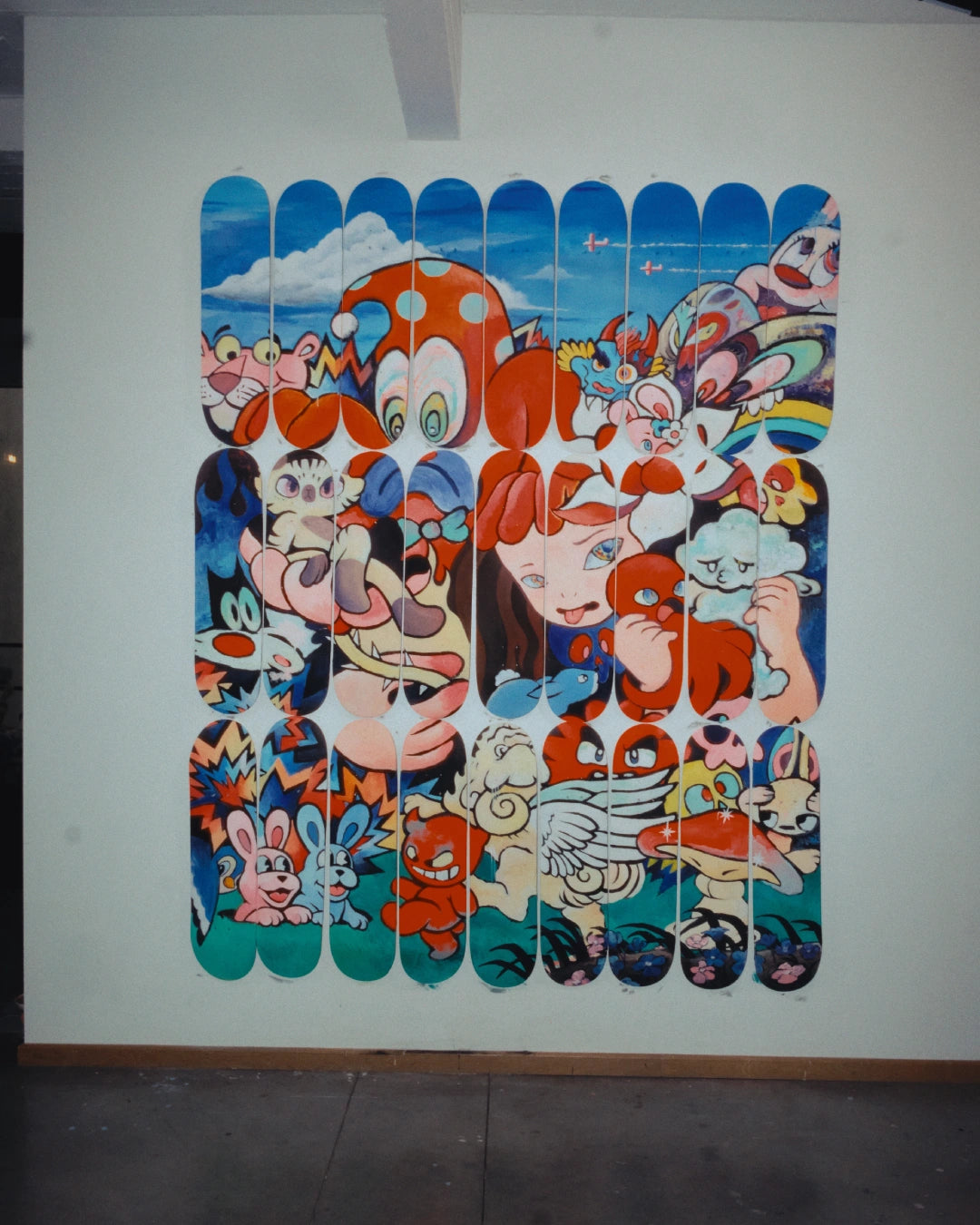 Full view of VEXX’s completed mural ‘The Life of the Party’ painted across 27 vertically mounted skateboard decks displayed on a studio wall.