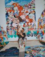 Artist VEXX smiling while holding a cat in front of his completed mural ‘The Life of the Party,’ surrounded by colorful original artworks and painted skate decks in his studio.