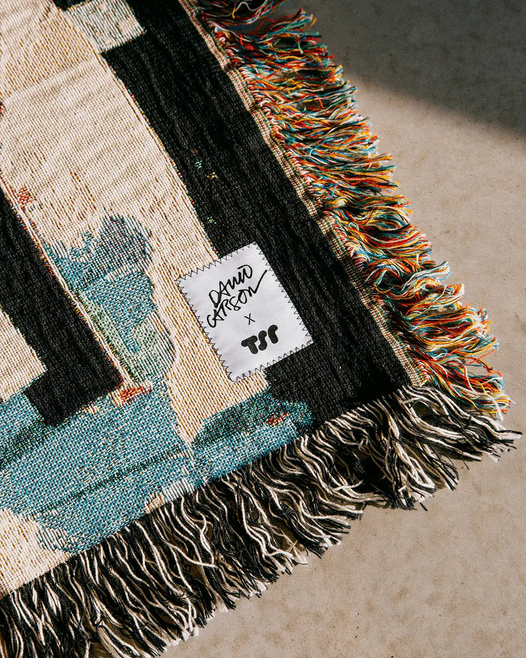 Multicolored woven blanket with a brand label saying David Carson x TSR on a neutral background