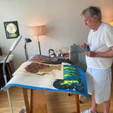 Artist Raymond Pettibon standing in his studio, working on a large-scale collaborative artwork with Marcel Dzama. The table is covered with vibrant, surreal prints featuring expressive figures and bold colors. Art supplies and sketches are scattered around, highlighting the creative process behind their limited-edition skate decks for THE SKATEROOM.