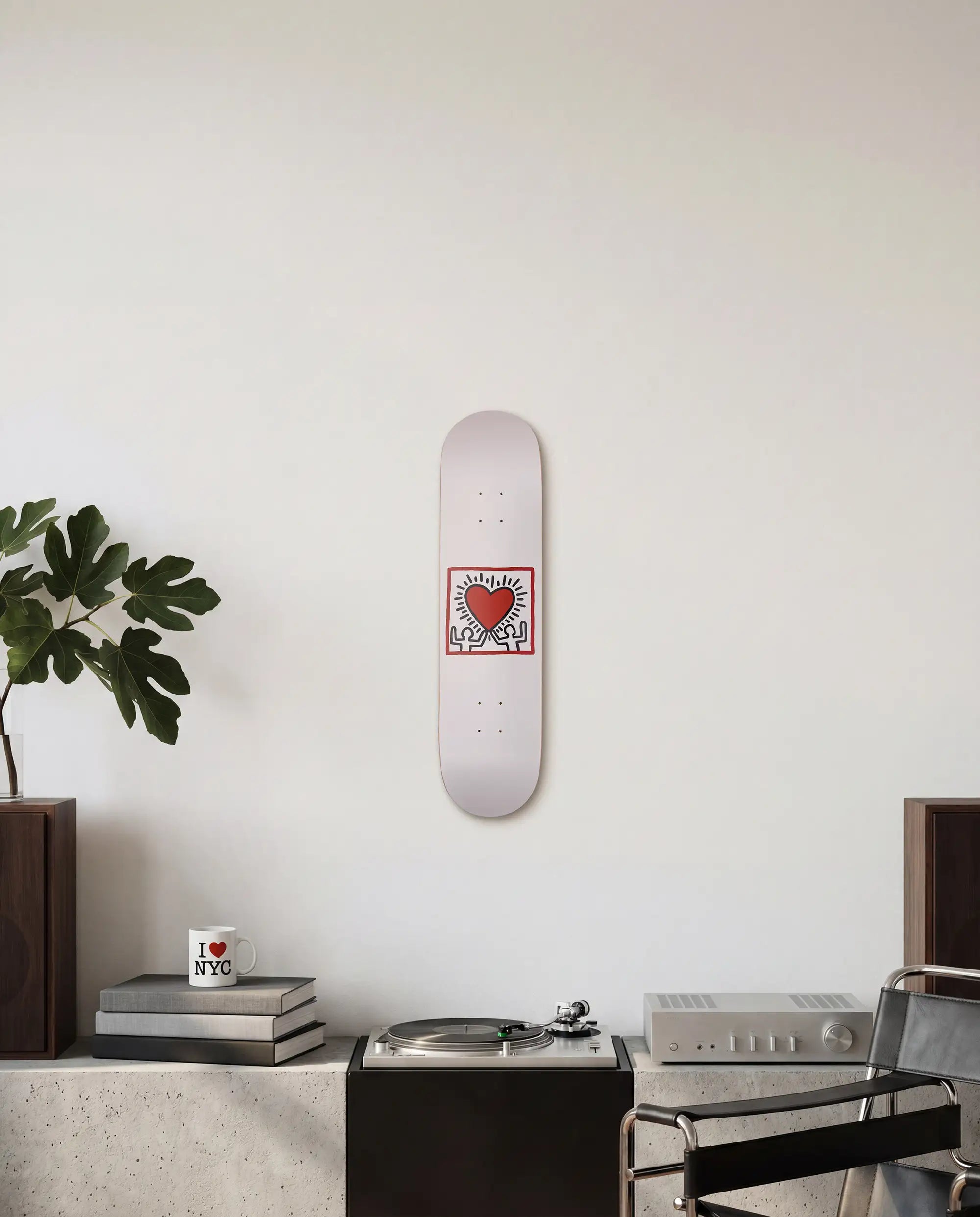 Skateboard deck with a radiant heart design by Keith Haring on a white wall above a table with books, a I LOVE NYC mug and a plant.
