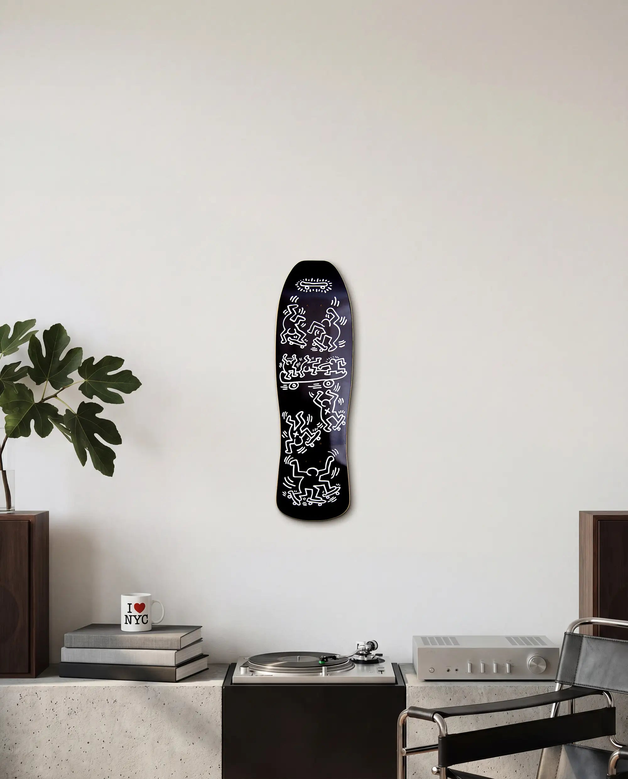 Black skateboard deck with white graphics by Keith Haring on a wall above a brutalist cupboard with books and a plant.