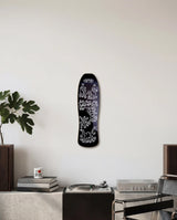 Black skateboard deck with white graphics by Keith Haring on a wall above a brutalist cupboard with books and a plant.