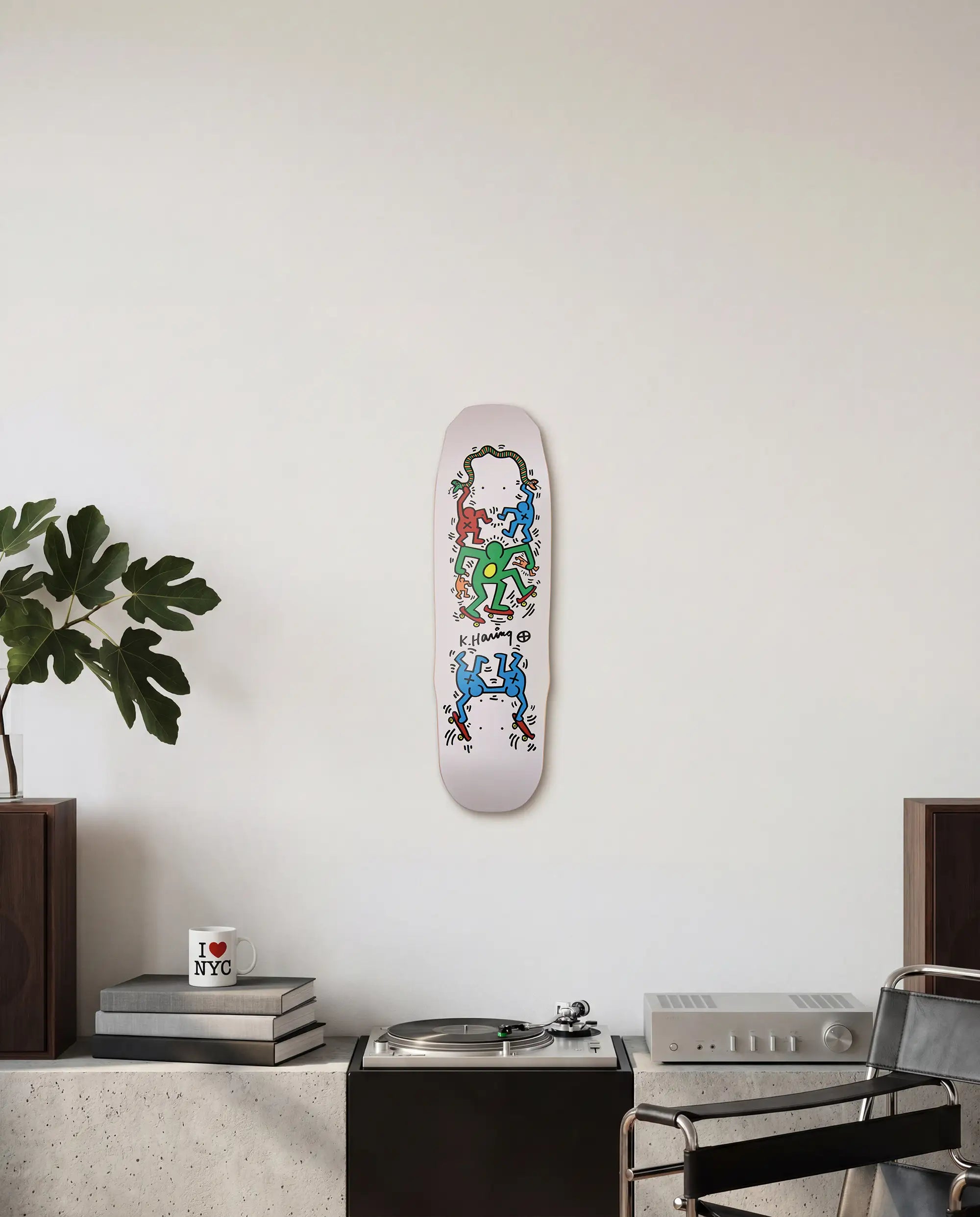 Skateboard deck with colorful Keith Haring design mounted as wall art on a white wall above a brutalist credenza with books and a plant.