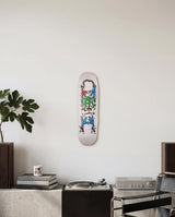 Skateboard deck with colorful Keith Haring design mounted as wall art on a white wall above a brutalist credenza with books and a plant.