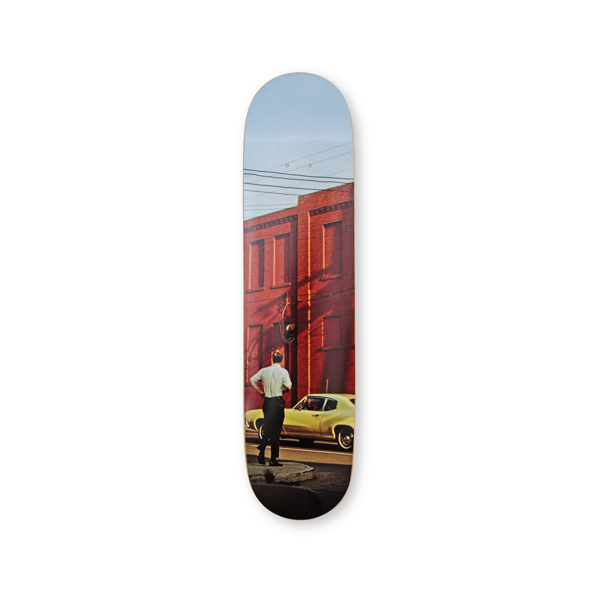 Skateboard deck by THE SKATEROOM and William Eggleston with a printed design of a man standing next to a yellow car in front of a red building.
